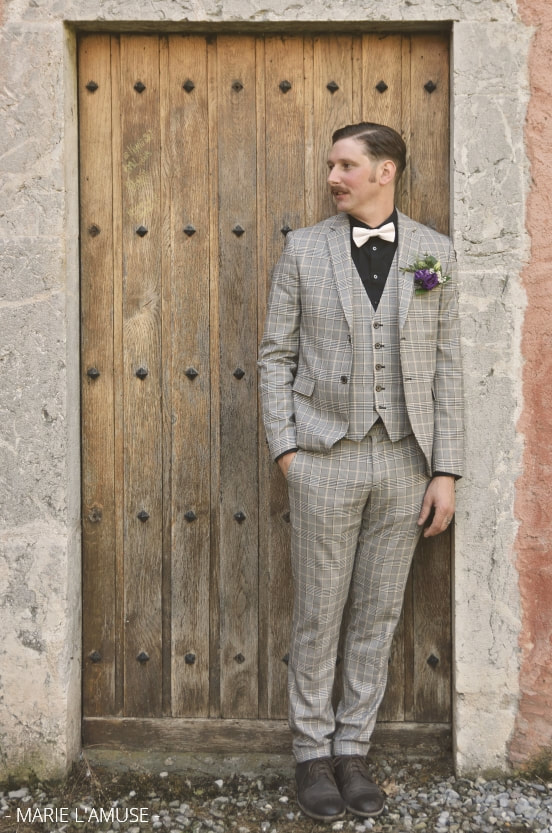 Mariage, Portrait, Le marié en costume à carreaux gris attend devant une porte en bois, Marignier Haute Savoie 2019, Photographe Marie l'Amuse
