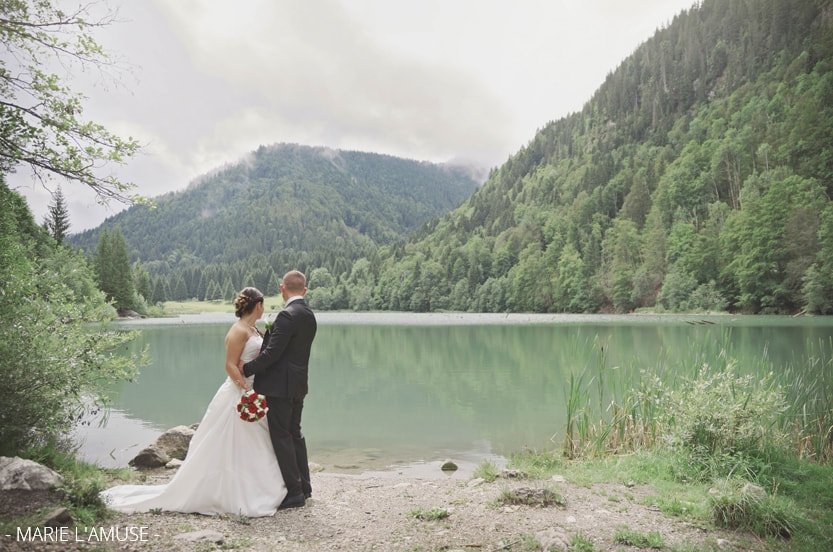 Séance photo de couple : ils regardent le lac de Vallon et la forêt, nuages sur les montagnes. Mariage, Bellevaux, Haute-Savoie.