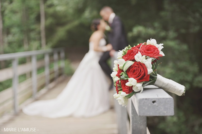 Séance photo couple : bouquet de roses rouges et blanc au premier plan, les mariés s'embrassent, flous, sur un pont. Mariage lac de Vallon Bellevaux, Haute-Savoie
