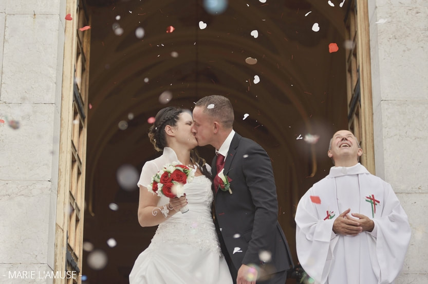 Mariage religieux, les mariés s'embrassent en sortant de l'église, le prêtre regarde le ciel, confettis flous. Habère-Poche Vallée Verte, Haute-Savoie